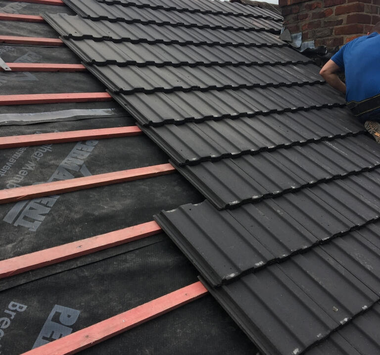 Roofer installing dark grey roof tiles over new battens and breathable membrane beside a brick chimney.